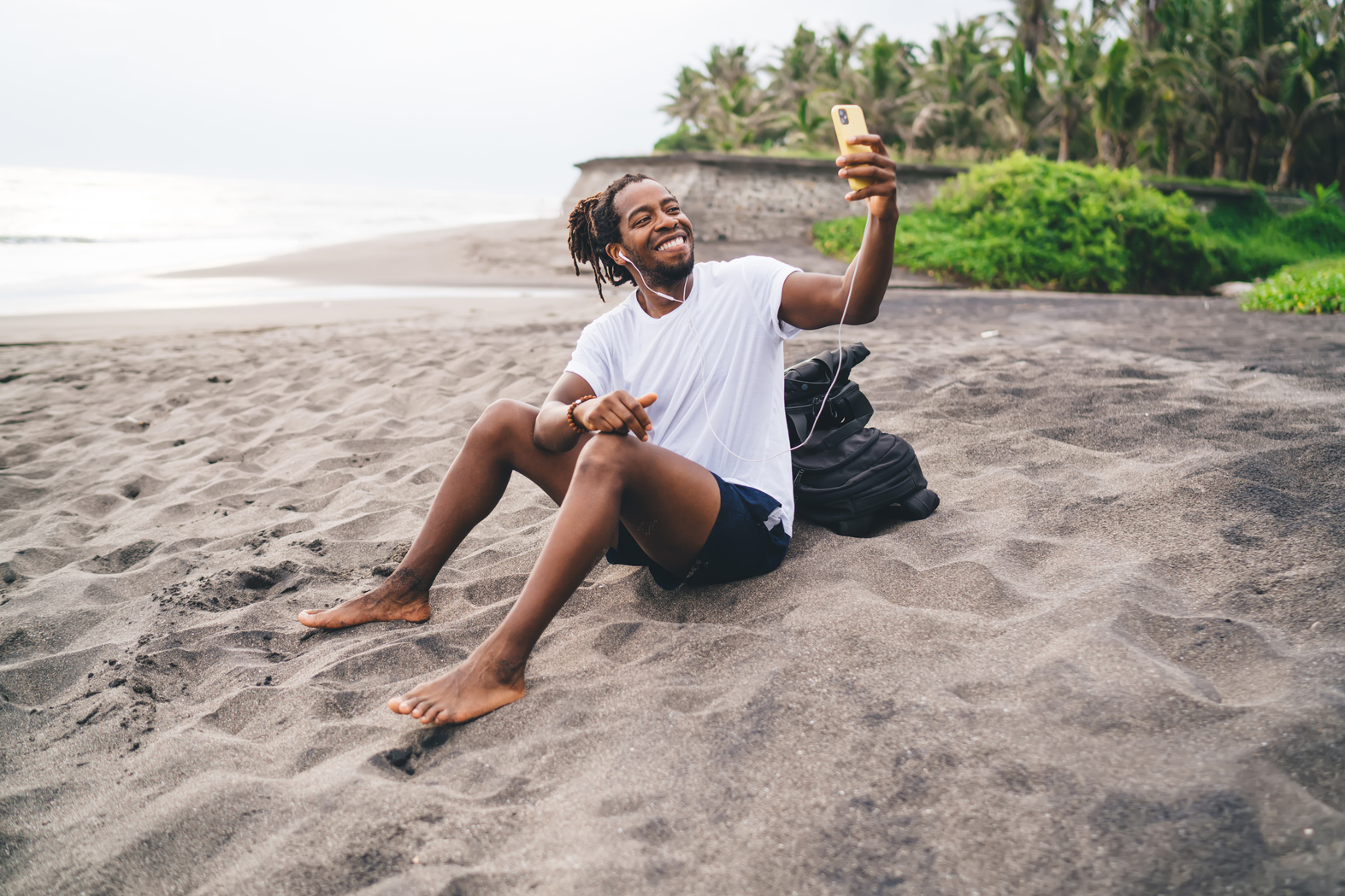 Happy ethnic man taking selfie on smartphone while sitting on beach